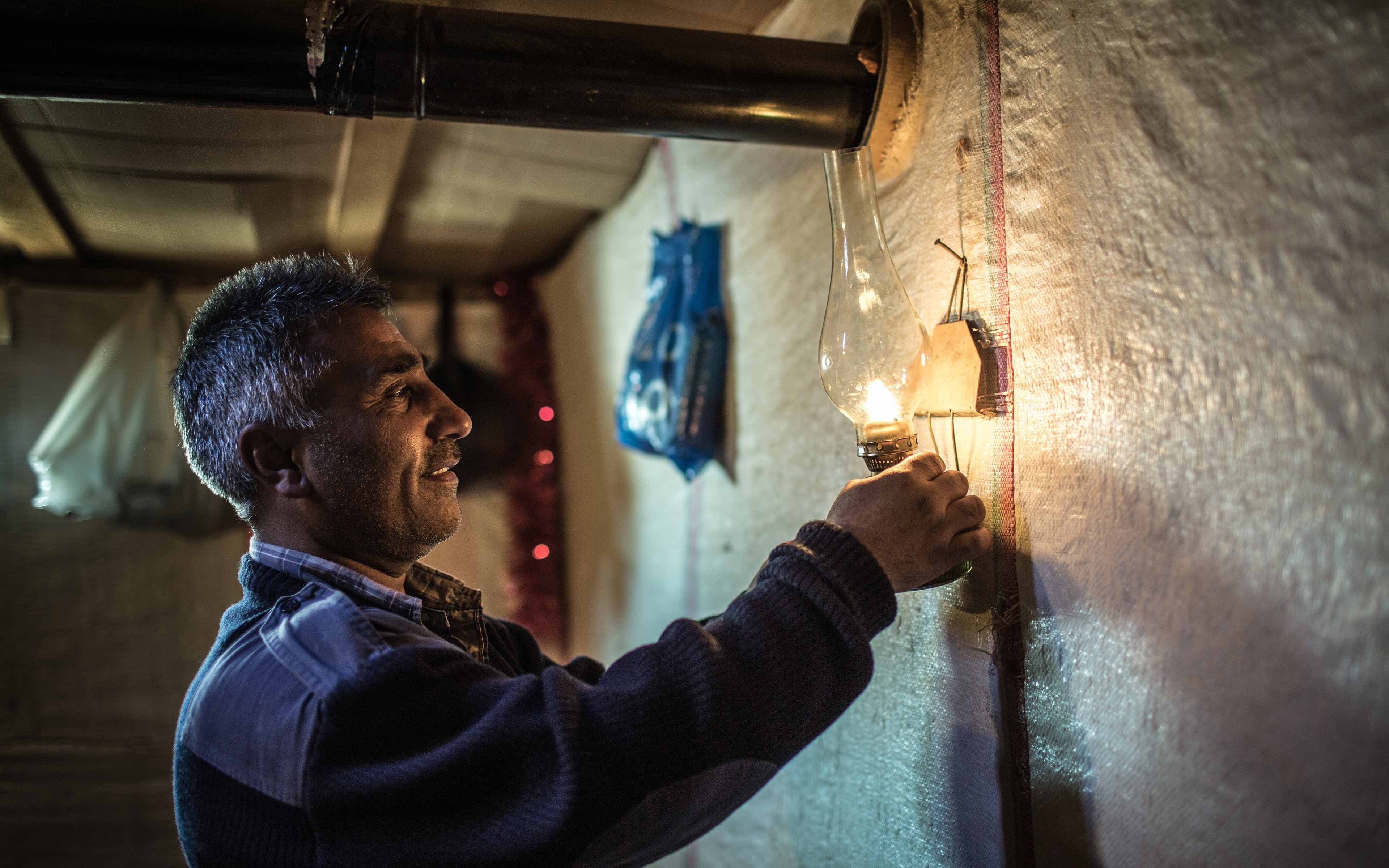 Abbas lights a kerosene lamp in a shelter made of plastic sheeting. He and his family fled from a suburb of Homs, Syria, and now live here in Lebanon. About 70 percent of Syrian refugees in Lebanon live below the national poverty line of $4 per person per day.